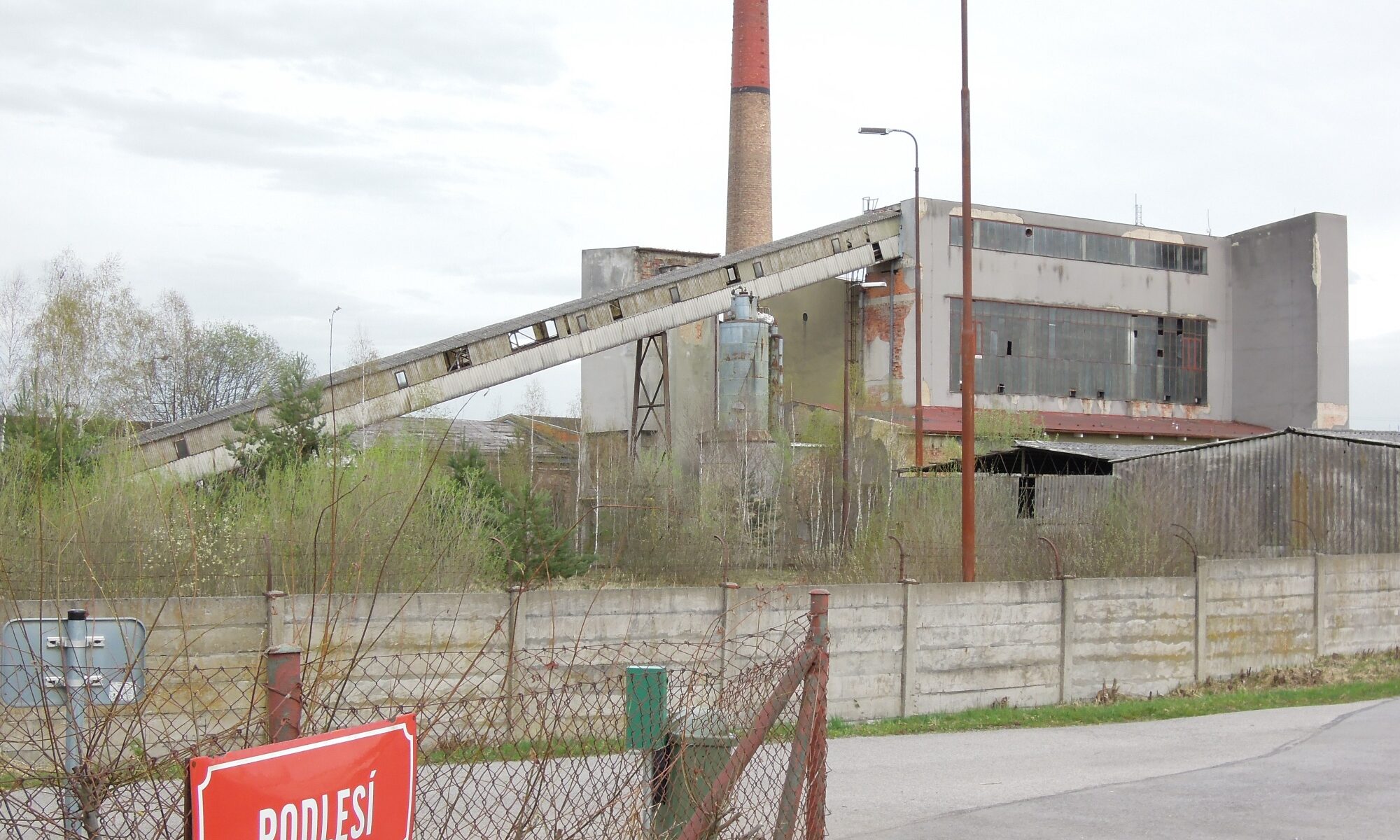 Foto aus dem schlot.at-Archiv (2024) zeigt Fabriksteil der Eisenbahnwerkstätten České Velenic in Tschechien, vermutlich Kraftwerk mit Schornstein und Schrägförderanlage für Kohle, im Vordergrund Maschendrahtzaun und rotes Schild mit Aufschrift "PODLESÍ"