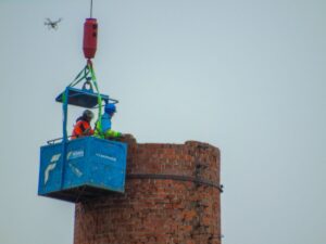 Schlot bei der Kürzung. Krankorb mit zwei Personen am oberen Ende des Schlots, Detail: oberhalb eine Drohne, die vermutlich den Abbruch filmt. Quelle: Sammlung Roland Barth